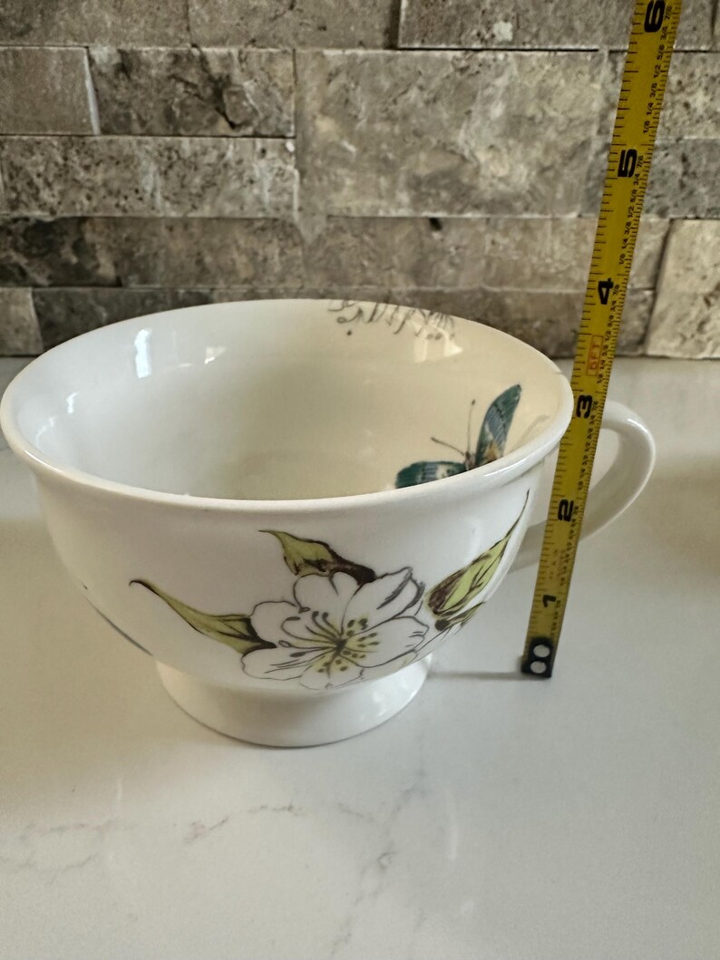 May include: White ceramic bowl with a handle, featuring a floral and butterfly design in green and blue. The bowl is approximately 4 inches in diameter and 3 inches tall. The bowl has a pedestal base.