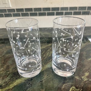 May include: Two clear glass tumblers featuring a white etched pattern of golf clubs, golf balls, and flags scattered across the exterior surface. The cylindrical drinking glasses have a smooth rim and a thick, weighted base for stability.
