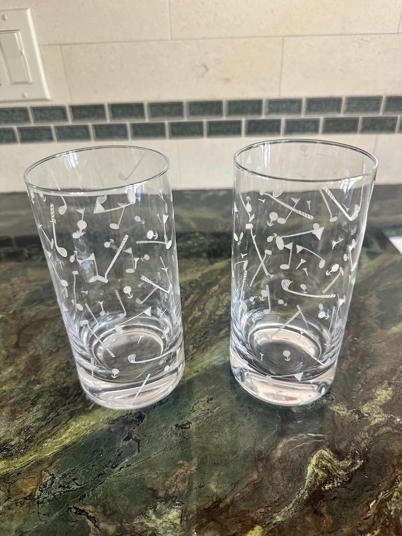 May include: Two clear glass tumblers featuring a white etched pattern of golf clubs, golf balls, and flags scattered across the exterior surface. The cylindrical drinking glasses have a smooth rim and a thick, weighted base for stability.