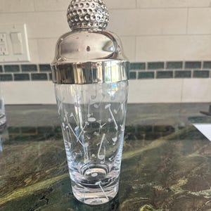 May include: A clear glass cocktail shaker featuring a white etched pattern of golf clubs and golf balls. The shaker is topped with a polished silver-toned metal lid shaped like a textured golf ball, providing a secure closure for the vessel.