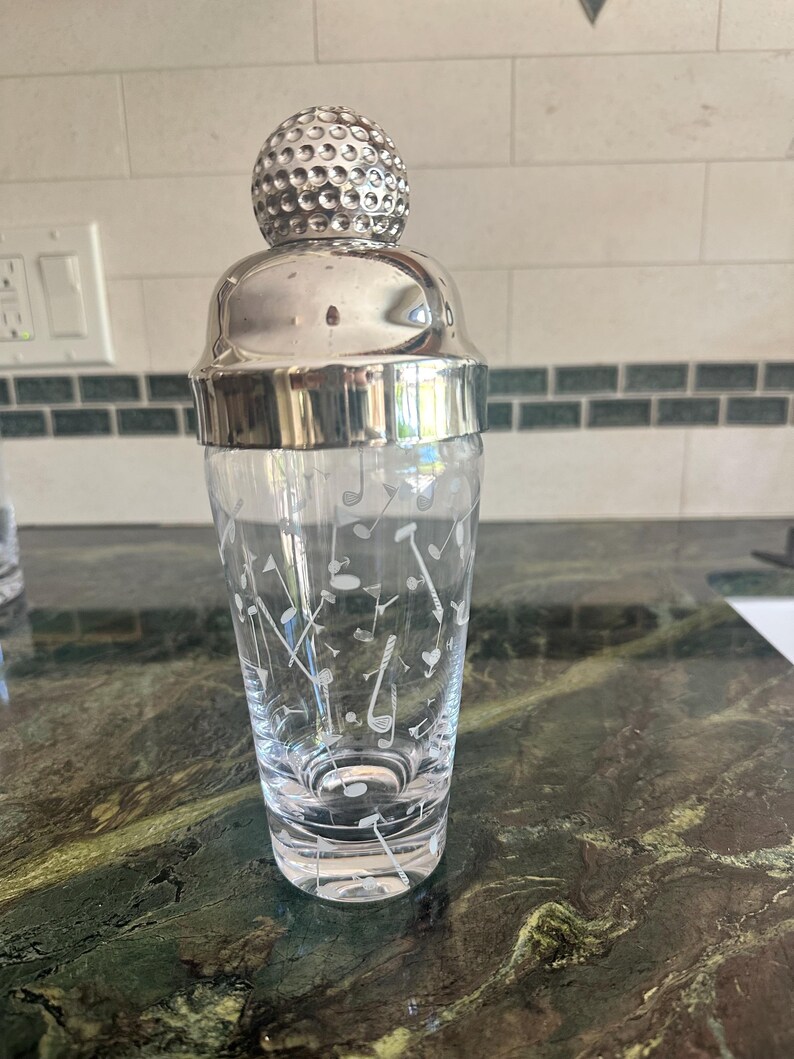 May include: A clear glass cocktail shaker featuring a white etched pattern of golf clubs and golf balls. The shaker is topped with a polished silver-toned metal lid shaped like a textured golf ball, providing a secure closure for the vessel.