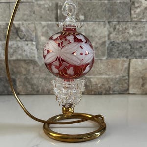 May include: A decorative glass ornament with a red base and clear floral design. The ornament hangs from a gold-colored metal stand. The ornament is suspended by a thin gold string.