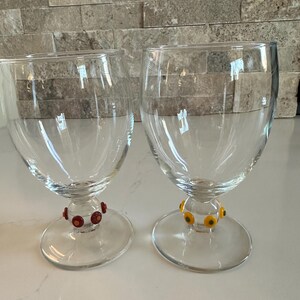 May include: Two clear stemmed wine glasses. One glass has a red and brown beaded design around the base of the stem. The other glass has a yellow and orange beaded design around the base of the stem.