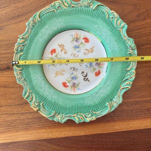 May include: A decorative plate with a mint green border and a floral center. The plate has a gold trim and measures approximately 8 inches in diameter. The center features a delicate floral pattern with red, blue, and purple flowers.