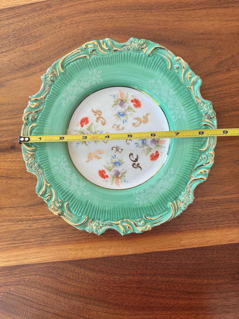 May include: A decorative plate with a mint green border and a floral center. The plate has a gold trim and measures approximately 8 inches in diameter. The center features a delicate floral pattern with red, blue, and purple flowers.