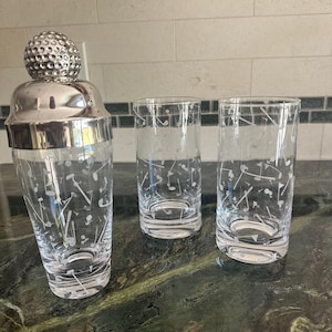May include: A glass cocktail shaker with a silver-toned lid shaped like a golf ball, accompanied by two matching tall drinking glasses. All three items feature a white etched pattern of golf clubs, golf balls, and flags across the clear glass.