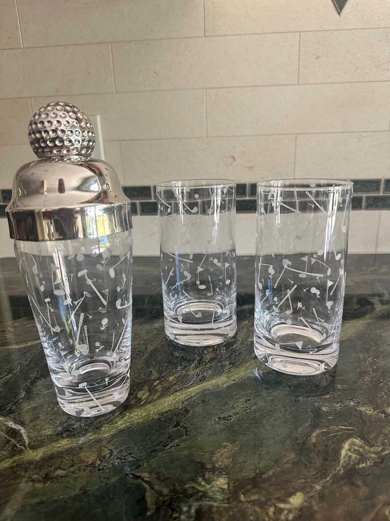 May include: A glass cocktail shaker with a silver-toned lid shaped like a golf ball, accompanied by two matching tall drinking glasses. All three items feature a white etched pattern of golf clubs, golf balls, and flags across the clear glass.