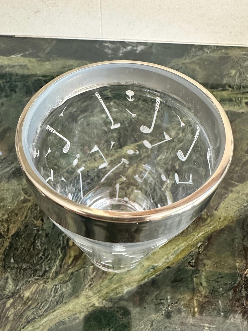 May include: A clear glass tumbler featuring a metallic silver rim and a white etched pattern of golf clubs, golf balls, and golf tees scattered across the exterior surface. The glass has a tapered cylindrical shape with a smooth, transparent finish.