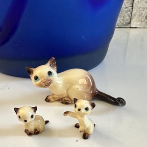 Op de afbeelding: Set van keramische Siamese kattenbeeldjes. De volwassen kat is crème en bruin, met blauwe ogen. Twee kleinere kittens zijn ook crème en bruin. De beeldjes staan op een wit oppervlak.