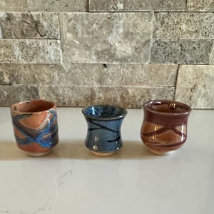 May include: Three handmade ceramic cups in a row. The cup on the left is orange with blue and white accents. The middle cup is blue with black lines. The cup on the right is brown with orange accents. All three cups have a light beige base.