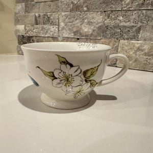May include: White ceramic teacup with a floral design featuring white flowers and green leaves. The teacup has a handle and sits on a small pedestal base. The background is a stone wall.