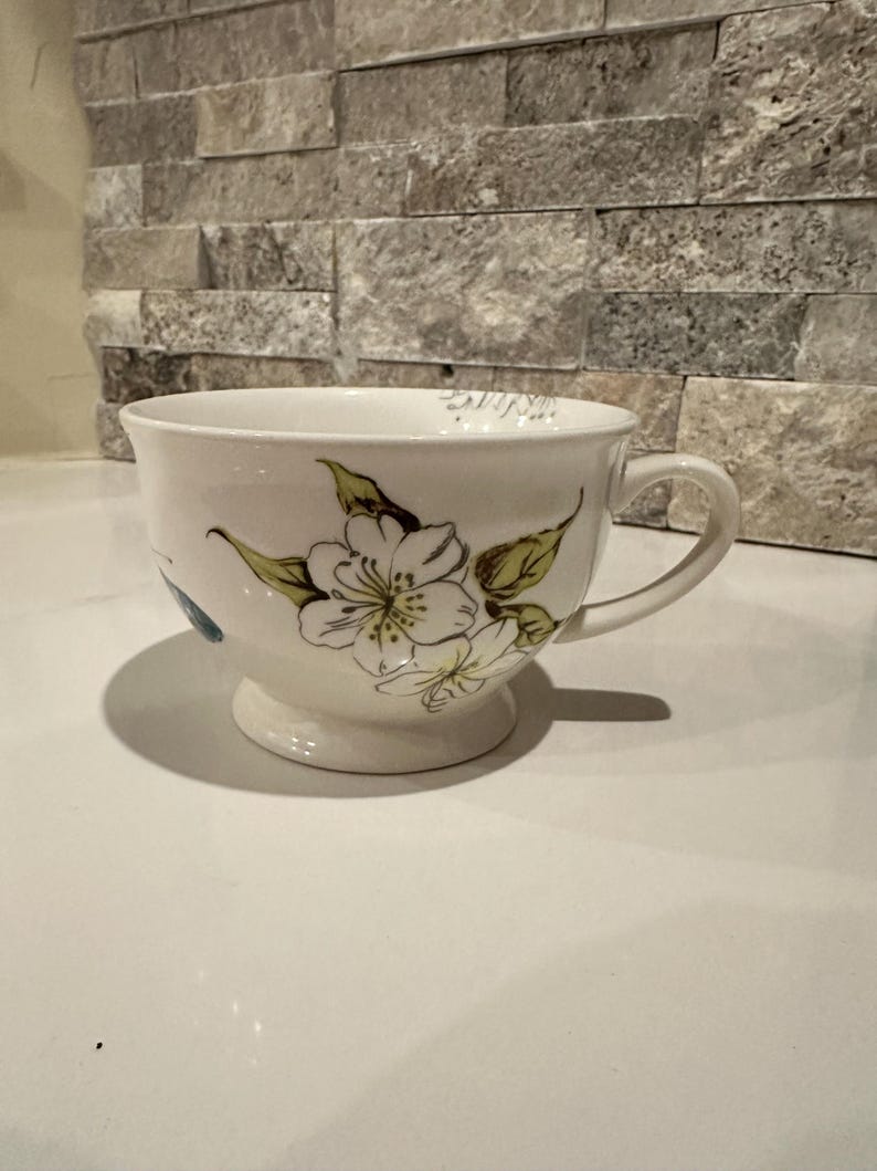 May include: White ceramic teacup with a floral design featuring white flowers and green leaves. The teacup has a handle and sits on a small pedestal base. The background is a stone wall.