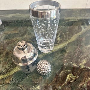 May include: A three-piece cocktail shaker set featuring a clear glass tumbler with a white etched golf-themed pattern, a silver-toned metal strainer lid, and a separate silver-toned metal cap shaped like a textured golf ball.