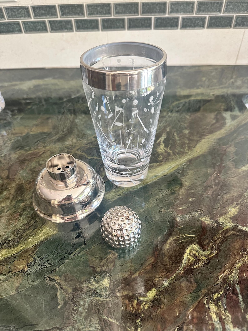 May include: A three-piece cocktail shaker set featuring a clear glass tumbler with a white etched golf-themed pattern, a silver-toned metal strainer lid, and a separate silver-toned metal cap shaped like a textured golf ball.