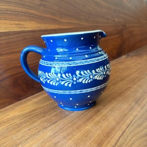 Può includere: Un'anfora in ceramica blu e bianca con un motivo floreale. L'anfora ha un manico bianco e un bordo bianco.