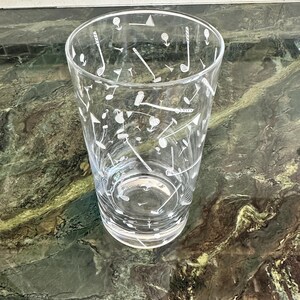 May include: A clear glass tumbler featuring a white etched pattern of golf-themed icons, including golf clubs, golf balls, tees, and flags scattered across the exterior surface. The glass has a cylindrical shape with a thick, solid base.