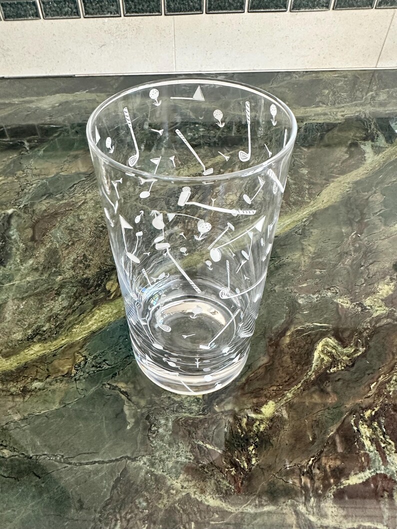 May include: A clear glass tumbler featuring a white etched pattern of golf-themed icons, including golf clubs, golf balls, tees, and flags scattered across the exterior surface. The glass has a cylindrical shape with a thick, solid base.