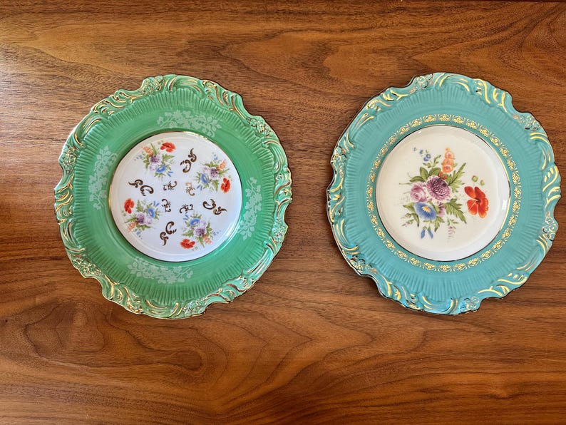 May include: Two decorative plates with floral designs. One plate is teal with a gold trim and a white center with a floral arrangement. The other plate is green with a gold trim and a white center with a floral design.