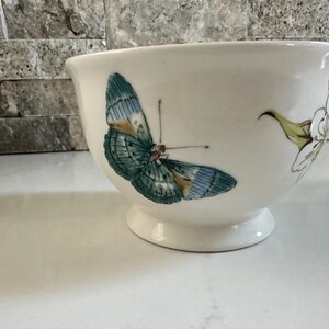 May include: Cream-colored ceramic bowl with a pedestal base, adorned with a detailed butterfly illustration in shades of teal, blue, and gold. A delicate floral design complements the butterfly. A decorative piece for the home.
