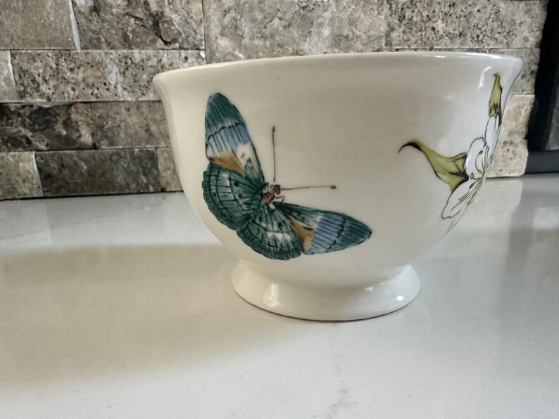 May include: Cream-colored ceramic bowl with a pedestal base, adorned with a detailed butterfly illustration in shades of teal, blue, and gold. A delicate floral design complements the butterfly. A decorative piece for the home.