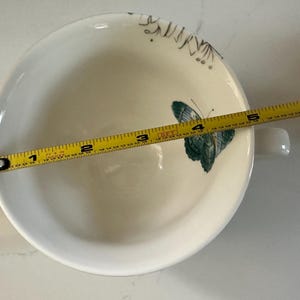 May include: White ceramic bowl with a handle, featuring a teal butterfly design. The bowl is approximately 6 inches in diameter. Delicate floral accents are also visible. A yellow measuring tape is placed across the bowl.