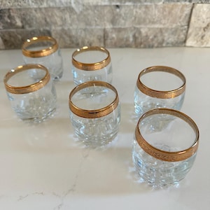 Könnte beinhalten: Sechs klare Glasbecher mit goldenem Rand. Die Gläser haben ein strukturiertes Muster auf der unteren Hälfte. Die Gläser sind auf einer weißen Oberfläche angeordnet.