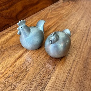 Puede incluir: Un juego de dos saleros y pimenteros de cerámica de color plateado con forma de pollos. Los saleros están sobre una superficie de madera.