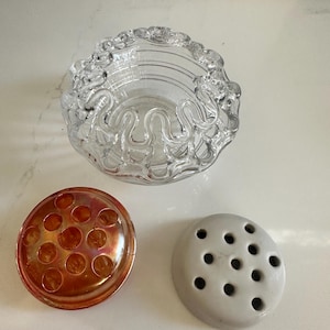 May include: A clear glass bowl with a wavy, scalloped rim, accompanied by two separate flower frog inserts. One insert is made of iridescent orange glass with circular holes, and the other is a solid, matte gray ceramic piece with holes.
