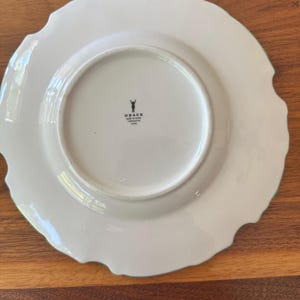 May include: White ceramic plate with a scalloped edge and a light green trim. The plate has a smooth, glossy finish and a circular indentation in the center. The word "GRACE" is printed on the bottom of the plate.