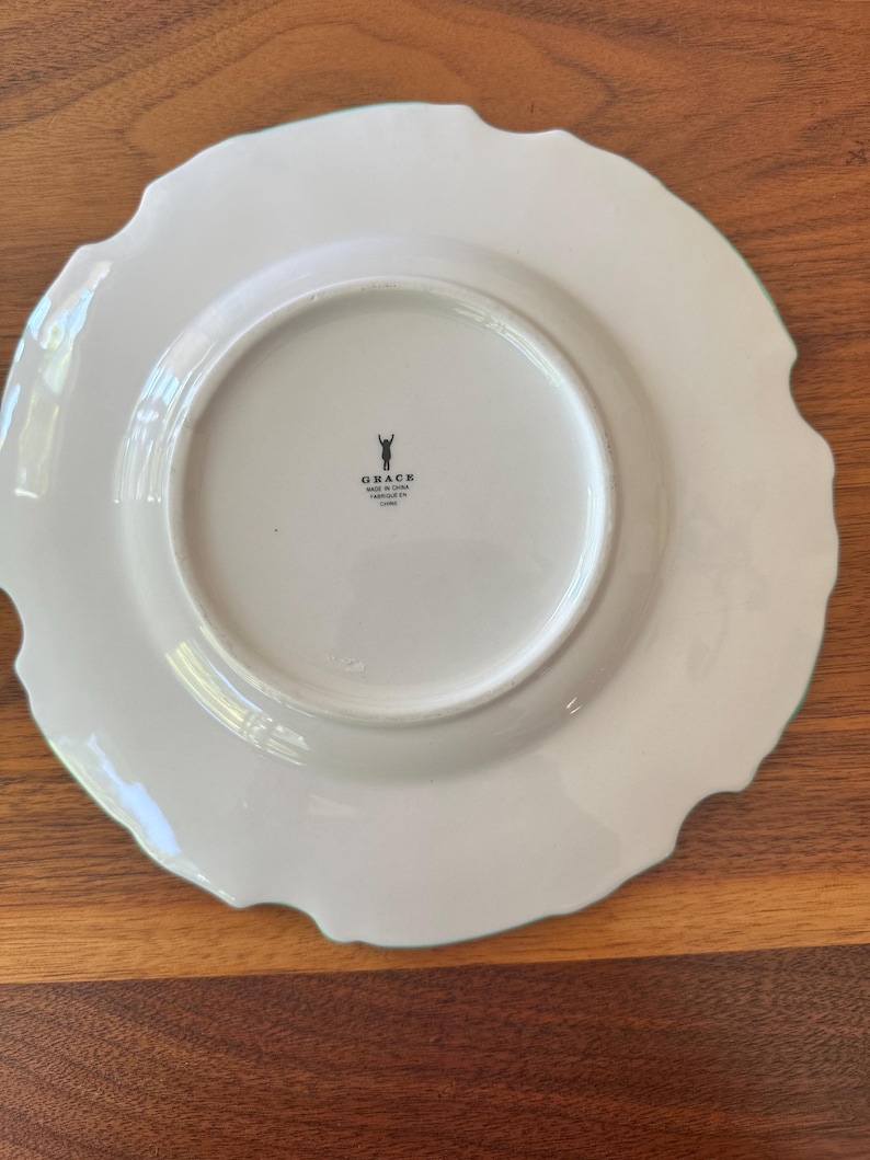May include: White ceramic plate with a scalloped edge and a light green trim. The plate has a smooth, glossy finish and a circular indentation in the center. The word "GRACE" is printed on the bottom of the plate.