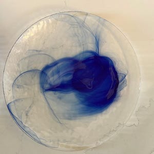 May include: A clear glass decorative plate with a vibrant blue abstract design. The plate features a central swirl of deep blue, with lighter blue wisps extending outwards. The plate has a slightly irregular edge.