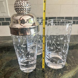 May include: A glass cocktail shaker with a silver-toned golf ball lid and two matching highball glasses, all featuring etched golf club and ball motifs. A yellow measuring tape shows the shaker stands approximately 10 inches in height.
