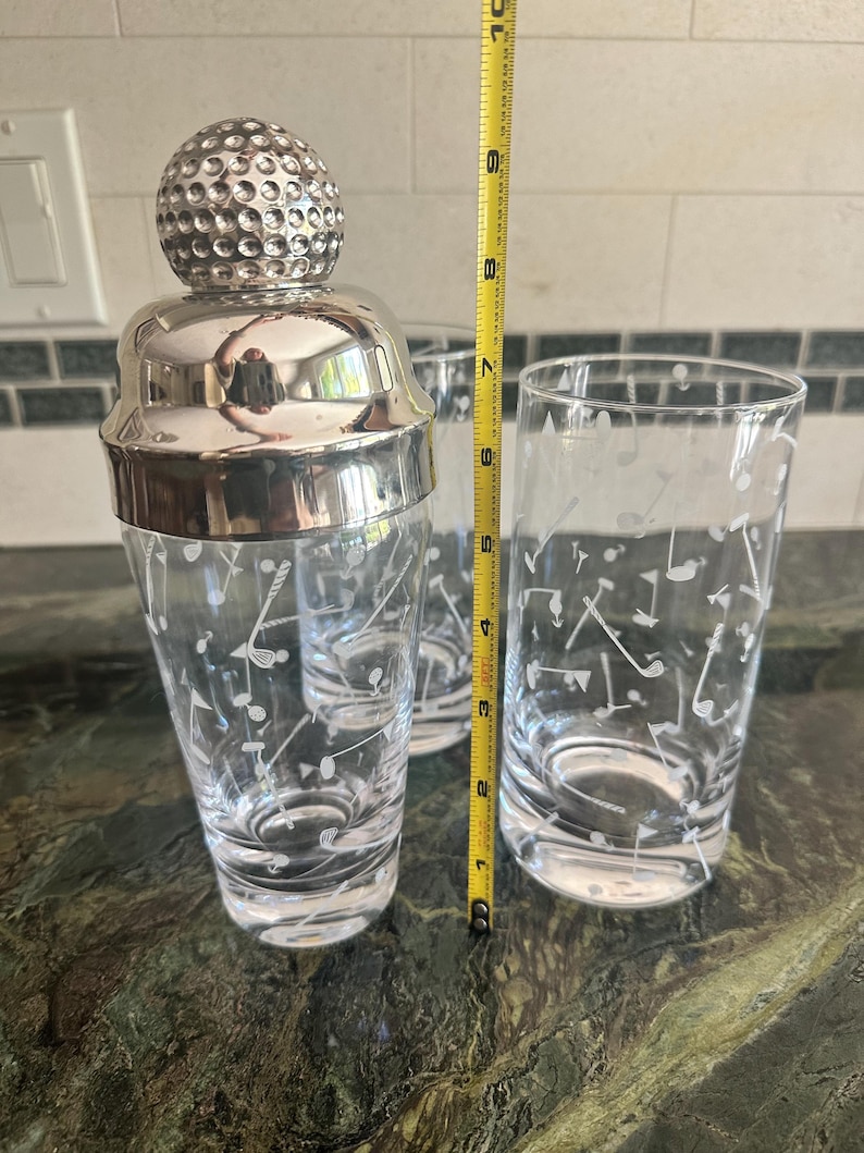 May include: A glass cocktail shaker with a silver-toned golf ball lid and two matching highball glasses, all featuring etched golf club and ball motifs. A yellow measuring tape shows the shaker stands approximately 10 inches in height.