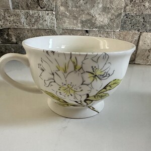 May include: White ceramic teacup with a floral design. The cup features a large handle and a pedestal base. The floral design includes white flowers with yellow centers and green leaves. The cup is approximately 4 inches in diameter.