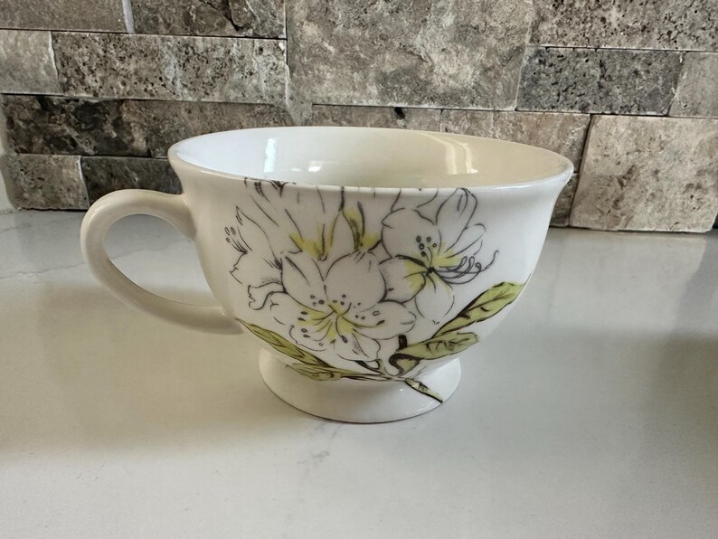 May include: White ceramic teacup with a floral design. The cup features a large handle and a pedestal base. The floral design includes white flowers with yellow centers and green leaves. The cup is approximately 4 inches in diameter.