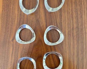 May include: Six silver napkin rings, each with a unique, slightly irregular, organic shape. The rings are arranged in two rows of three on a wooden surface.