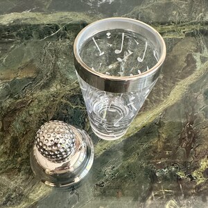 May include: A clear glass cocktail shaker featuring a white golf-themed pattern of clubs and tees, topped with a silver-toned lid textured to resemble a golf ball. The shaker includes a smooth silver-colored rim around the top opening.
