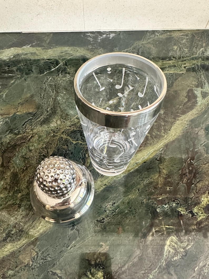 May include: A clear glass cocktail shaker featuring a white golf-themed pattern of clubs and tees, topped with a silver-toned lid textured to resemble a golf ball. The shaker includes a smooth silver-colored rim around the top opening.