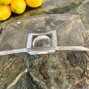 May include: A clear, rectangular glass serving dish with a curved design. The dish features a central, spherical accent and is positioned on a marble countertop. Several yellow lemons are visible in the background.