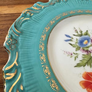 May include: Close-up of a decorative plate with a turquoise rim and a white center. The rim features gold accents and a scalloped edge. The center displays a floral design with blue, purple, and orange flowers.
