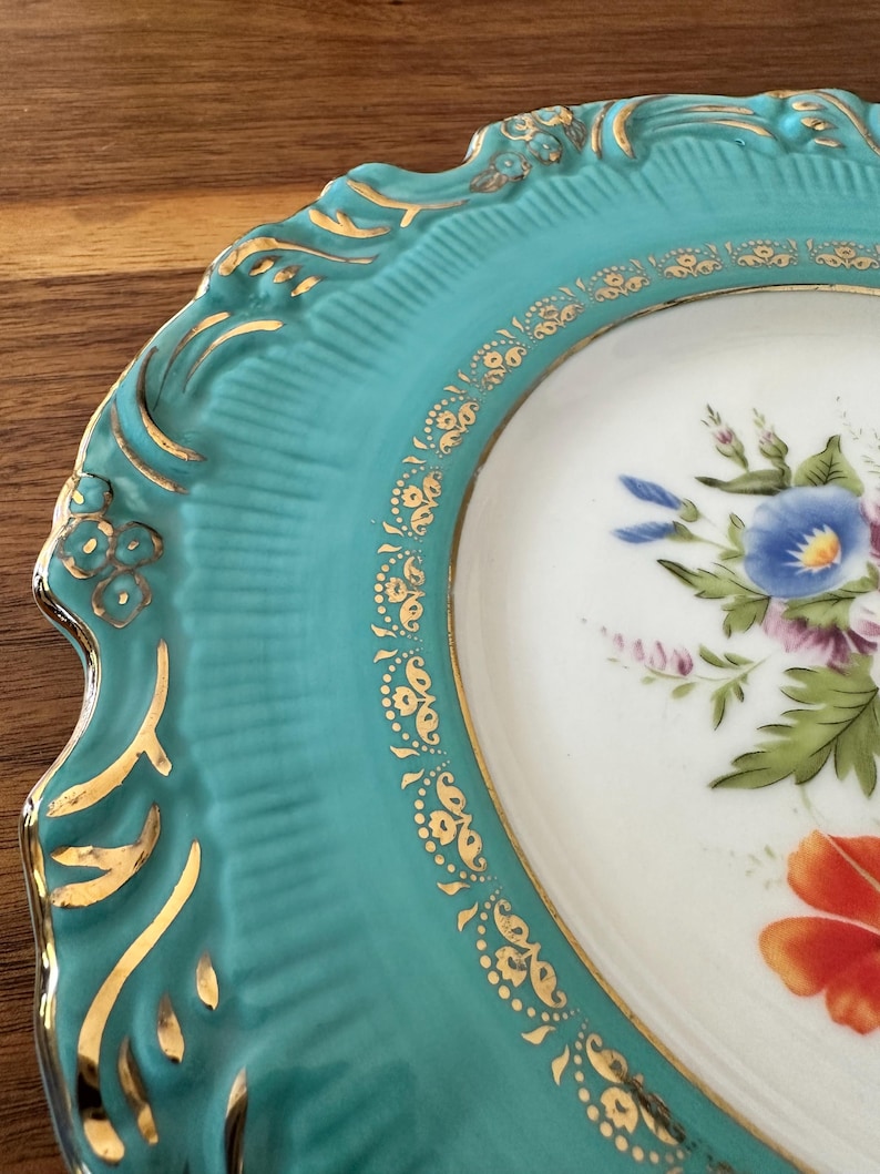 May include: Close-up of a decorative plate with a turquoise rim and a white center. The rim features gold accents and a scalloped edge. The center displays a floral design with blue, purple, and orange flowers.