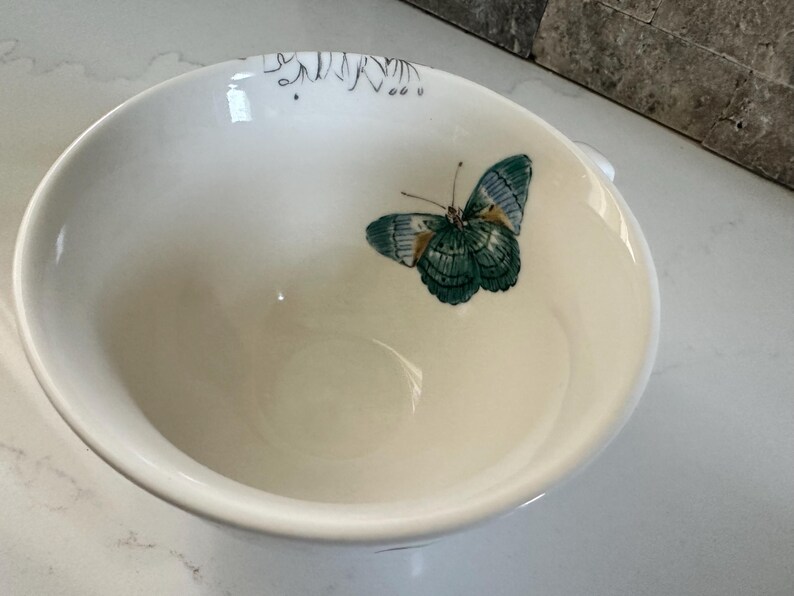 May include: White ceramic bowl with a butterfly design. The butterfly has teal and blue wings with a detailed pattern. The bowl has a smooth, glossy finish and a simple, elegant design.