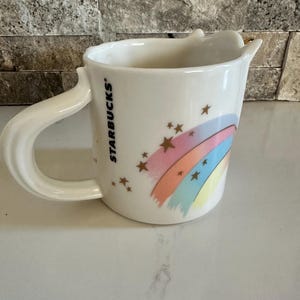 Peut inclure: Tasse en céramique blanche avec une anse incurvée, ornée d'un arc-en-ciel et d'étoiles. Le logo Starbucks est imprimé verticalement sur le côté. L'arc-en-ciel est composé de rose, orange, jaune et bleu.