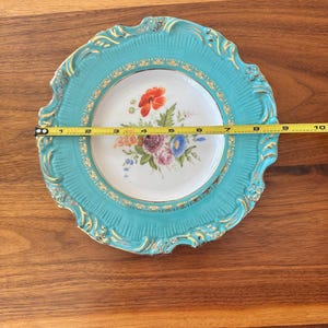 May include: A decorative plate with a turquoise rim and a floral design. The plate measures approximately 9 inches in diameter. The rim has a gold trim and ornate detailing. The center of the plate features a bouquet of colorful flowers.