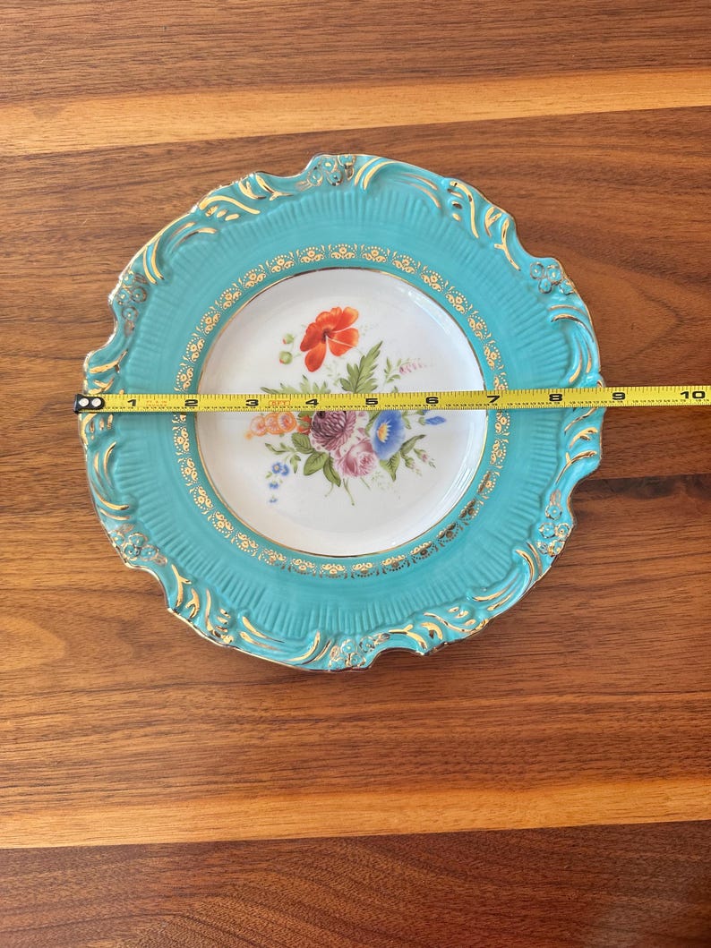 May include: A decorative plate with a turquoise rim and a floral design. The plate measures approximately 9 inches in diameter. The rim has a gold trim and ornate detailing. The center of the plate features a bouquet of colorful flowers.