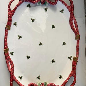May include: Oval white ceramic serving platter with a red rope-like border and holly accents. The border has gold accents and bow details at the top and bottom. Small holly sprigs are scattered across the white surface.