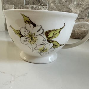 May include: White ceramic teacup with a floral design. The cup features a large, rounded bowl shape with a handle and a small pedestal base. The floral design includes white flowers and green leaves.