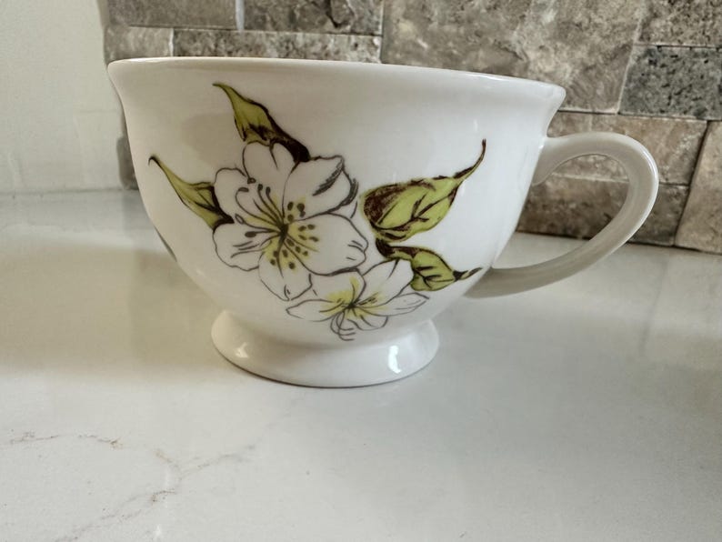 May include: White ceramic teacup with a floral design. The cup features a large, rounded bowl shape with a handle and a small pedestal base. The floral design includes white flowers and green leaves.