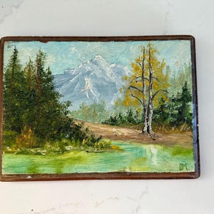 Puede incluir: Pintura al óleo de un paisaje de montaña con un lago y árboles. La pintura presenta una montaña nevada, un árbol amarillo y árboles de hoja perenne. La obra de arte está enmarcada en un marco de madera marrón oscuro.