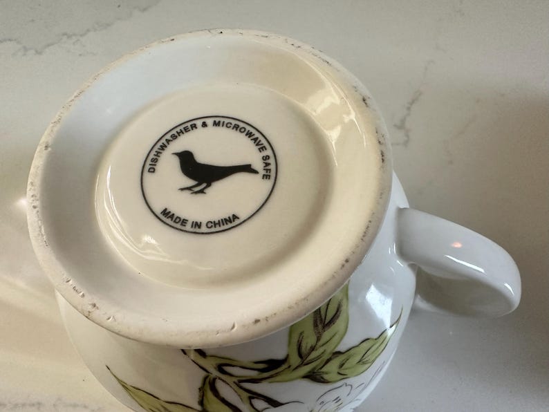 May include: White ceramic mug with a floral design and a handle. The bottom of the mug features a black bird silhouette inside a circle with the text "Dishwasher & Microwave Safe" and "Made in China".